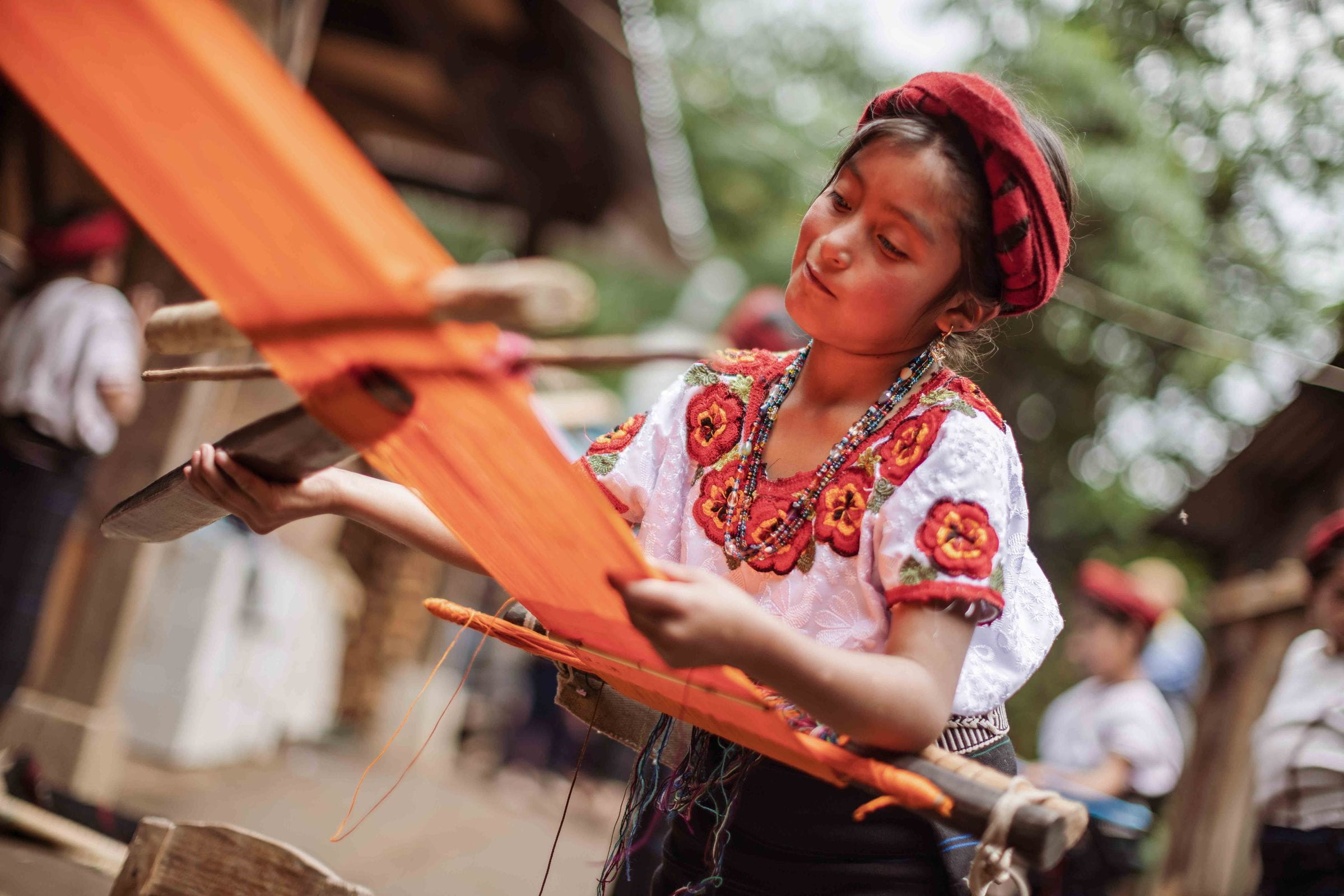 Ein Mädchen in traditioneller Kleidung webt mit orangefarbenem Garn unter freiem Himmel.  (Quelle: Jakob Studnar)